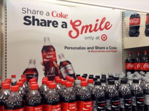 A shelf full of Coca-Cola's soda bottles that have people's names on them. The sign on the wall reads "Share a Coke. Share a Smile"